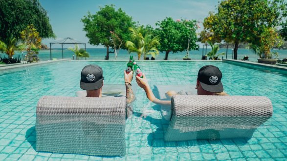 two men sat in pool bed with black hats on cheersing with beers
