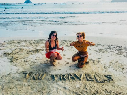Two girls on a sandy beach in Bali, Indonesia with TruTravels carved into the sand 