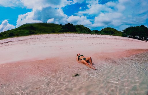 A fish eye lens shot of a girl laying on Pink Beach in Indonesia showing the pink sand, clear water and cloudy blue sky 